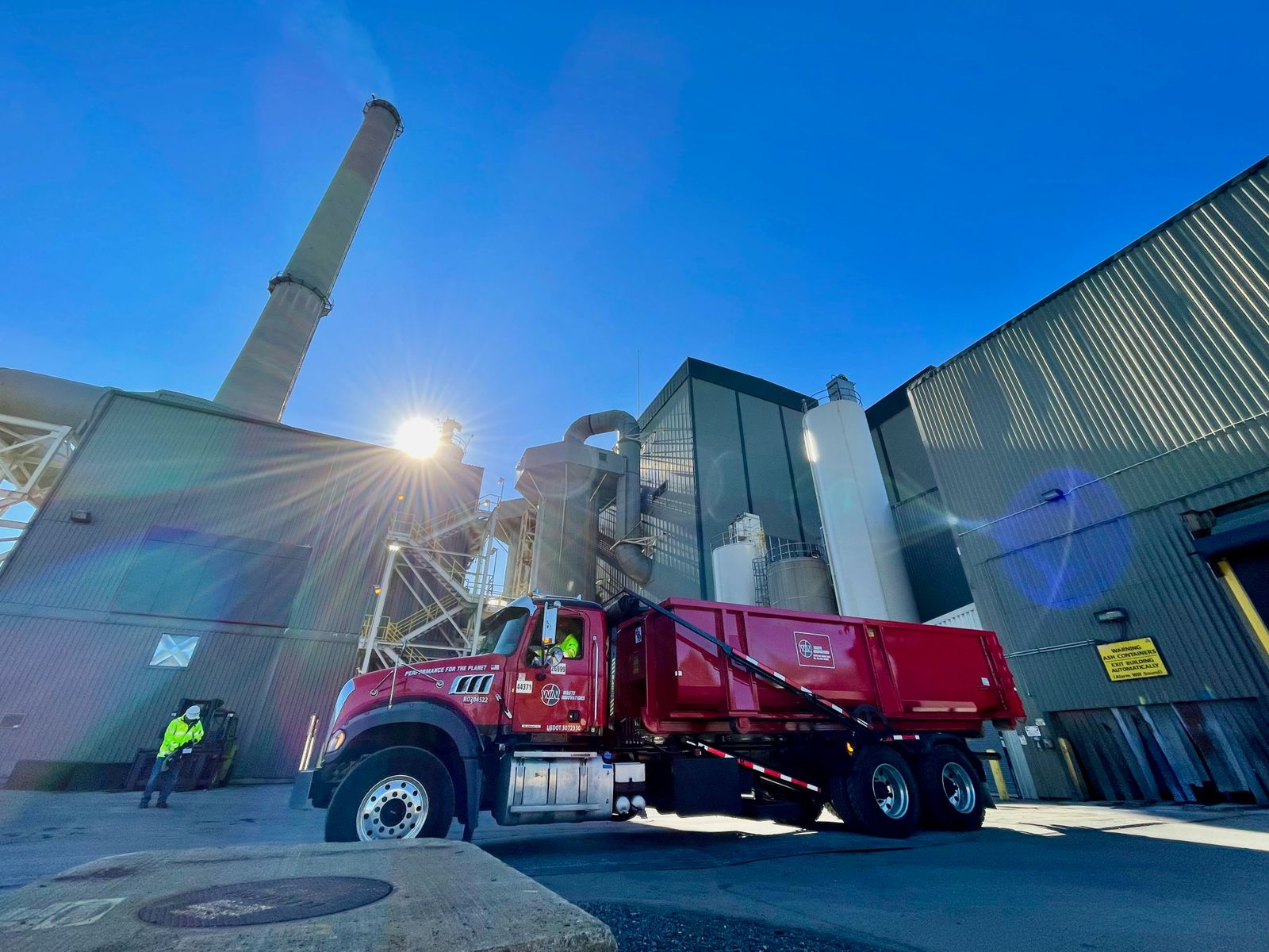 Red waste truck in front of industrial facility with smokestack in the background